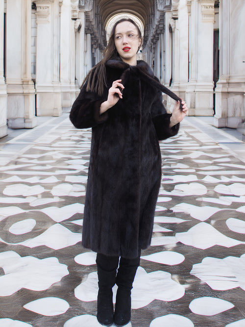 Model wearing La Galleria full-length mahogany mink fur coat with tie closure, styled in a grand architectural setting for winter fashion.