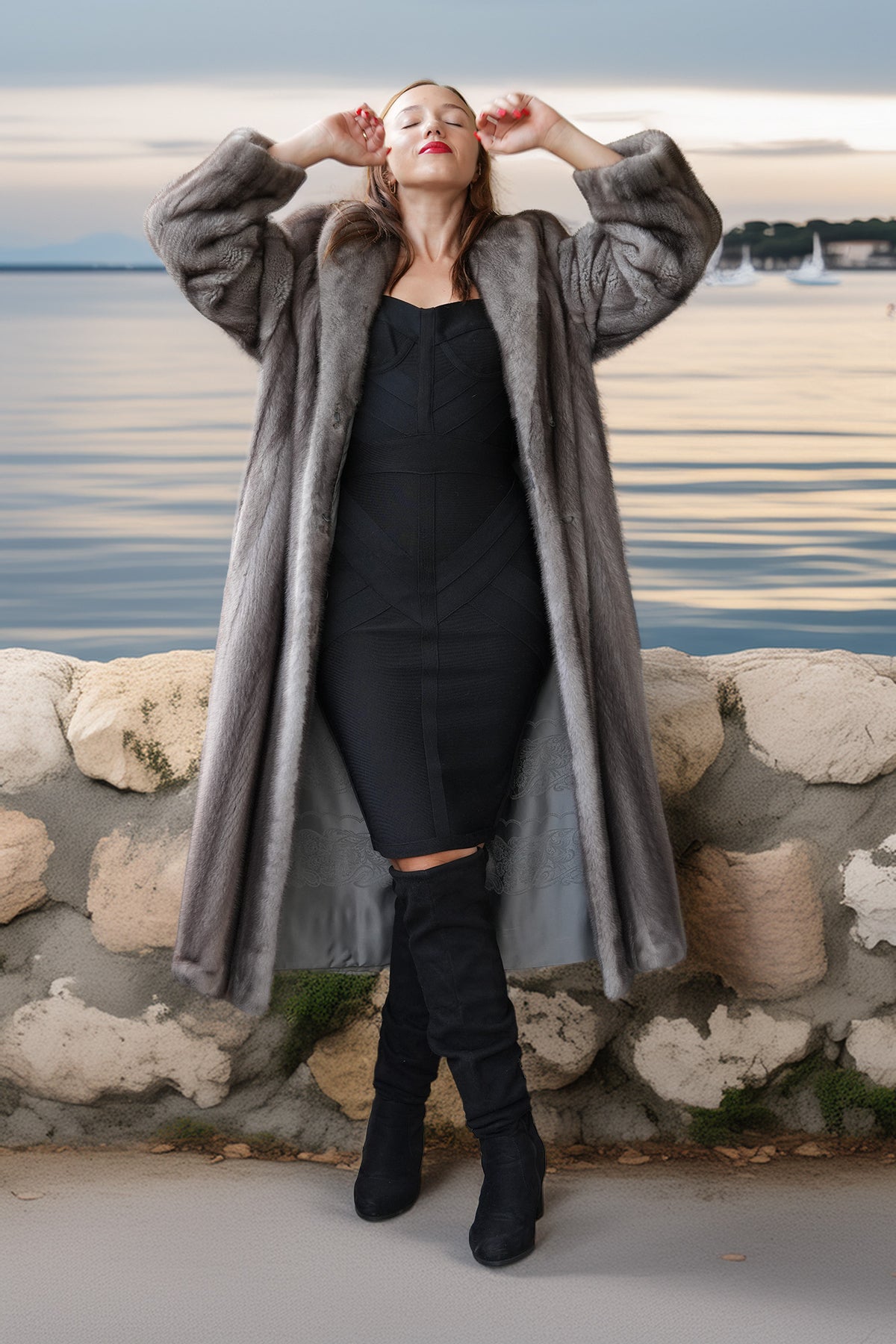 A woman wearing a long gray faux fur coat over a black dress and black knee-high boots, posing outdoors in front of a stone wall with water and a cloudy sky in the background.
