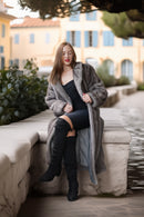 A woman sits on a stone ledge outdoors wearing a long gray faux-fur coat over a black dress and black over-the-knee boots, with a blurred building and water in the background.