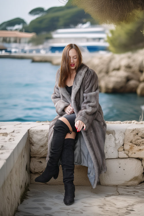 A woman sits on a stone ledge by the water wearing a long gray faux fur coat, black thigh-high boots, and a black top. The background shows a coastal marina with boats and green trees.