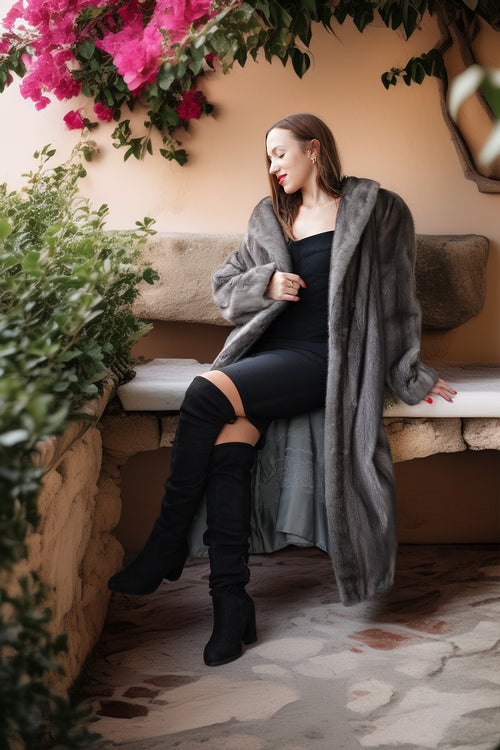A woman sits on a stone bench wearing a long gray faux fur coat over a black dress and black over-the-knee boots, with pink flowers and green plants in the background.
