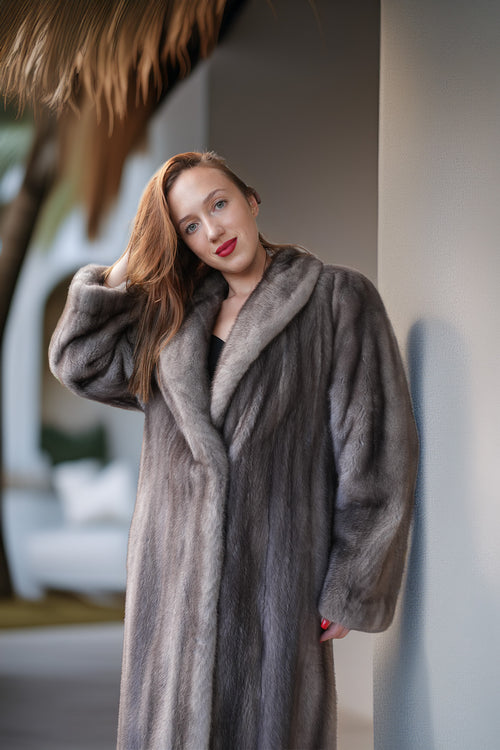 A woman wearing a luxurious, long gray fur coat with a wide collar, posing in a softly lit indoor setting with a thatched roof visible in the background.