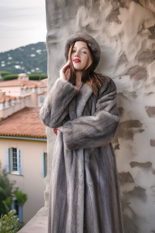A woman wearing a luxurious gray fur coat and matching fur hat leans against a textured stone wall, with a scenic Mediterranean village in the background.