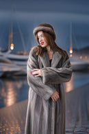 A woman wearing a luxurious gray fur coat and matching fur hat stands on a waterfront promenade at dusk, with boats and marina lights blurred in the background.