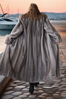 A woman viewed from behind wearing a long, voluminous gray fur coat while walking on a stone pier by the water at sunset.