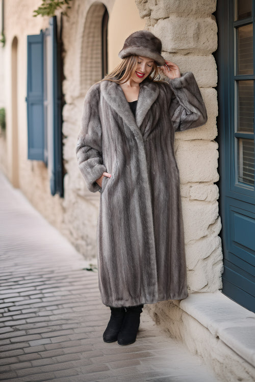 A woman wearing a long gray fur coat and matching fur hat stands on a cobblestone street beside a stone building with blue shutters.
