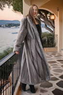 A woman wearing a long, luxurious gray fur coat over a black dress and black boots, standing on a stone walkway of a coastal terrace at sunset.