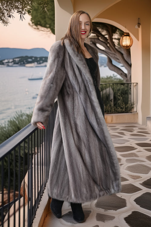 A woman wearing a long, luxurious gray fur coat over a black dress and black boots, standing on a stone walkway of a coastal terrace at sunset.