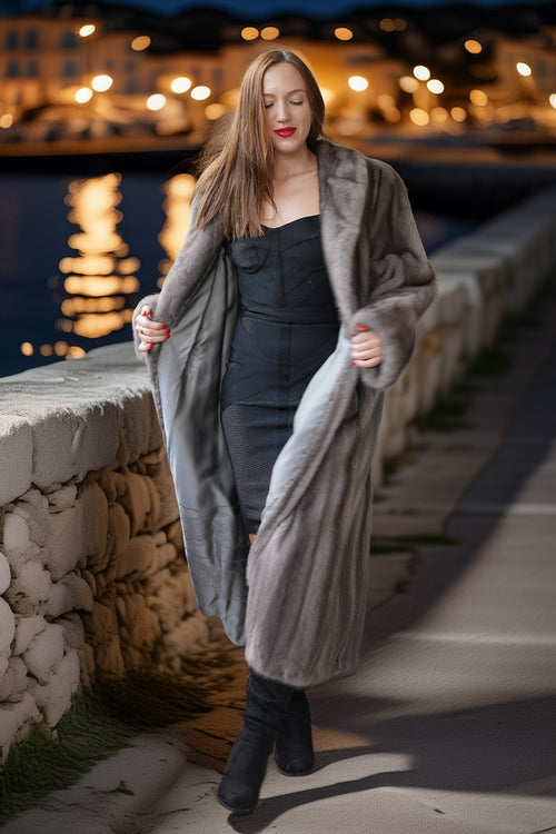 A woman wearing a long, plush gray fur coat over a black strapless dress and black boots, standing on a stone walkway at night with warm bokeh lights and water in the background.