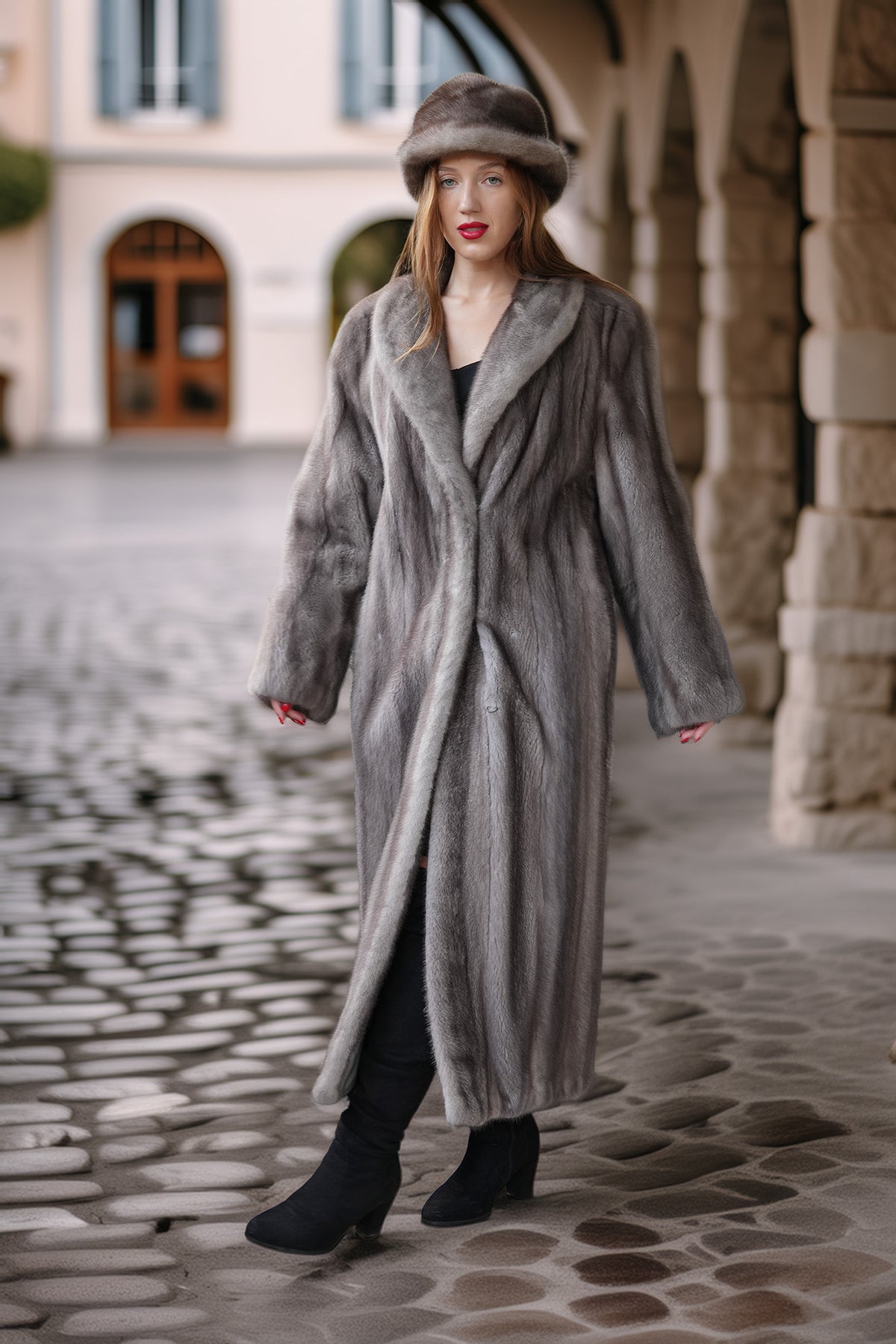 A woman wearing a long gray fur coat and matching fur hat stands on a cobblestone street in front of a stone archway building.