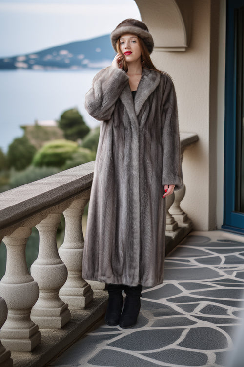 A woman stands on an outdoor balcony wearing a long gray fur coat, matching gray fur hat, black boots, and red lipstick, with a scenic lake and greenery in the background.
