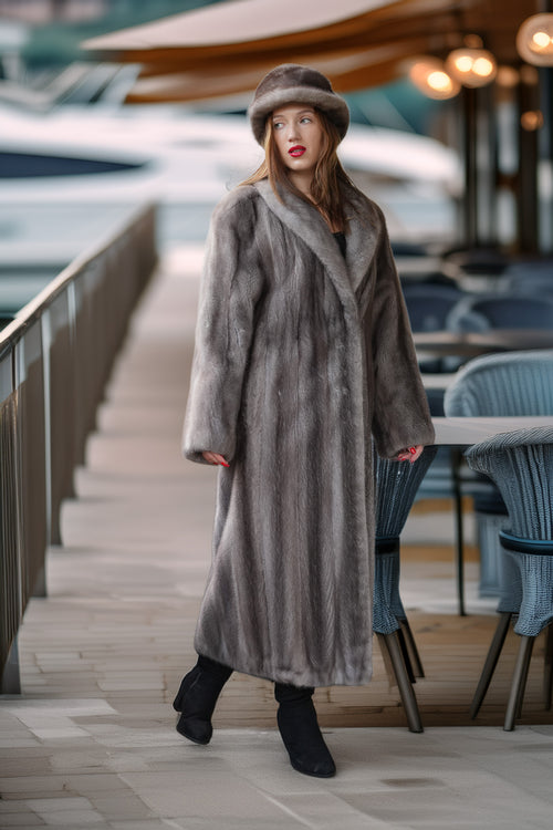 A woman models a long, luxurious gray fur coat and matching fur hat on an outdoor deck next to water, with a blurred background of boats and patio seating.
