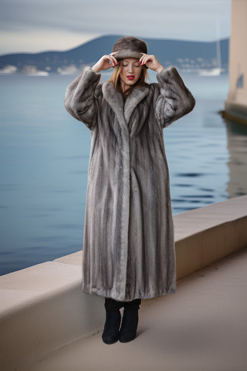 A woman wearing a long gray fur coat, matching fur hat, and black boots stands on a waterfront promenade with water and distant hills in the background.