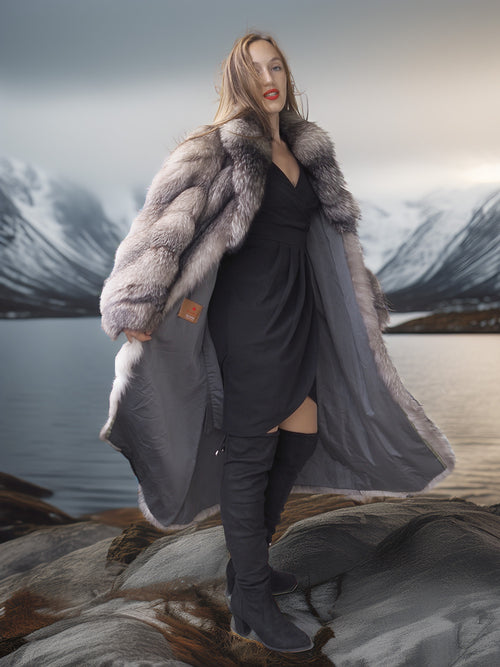 Woman in a long coat with fur trim standing on rocks by a lake with mountains in the background
