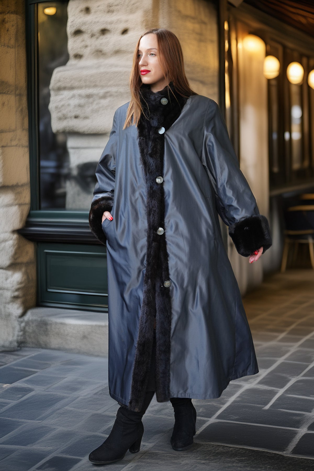 A woman wearing a long charcoal gray coat with black faux-fur trim and buttons, standing on a stone sidewalk in front of a building.