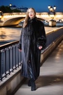 A woman wearing a long black fur coat and black boots walks along a bridge at night with a lit river bridge and water in the background.