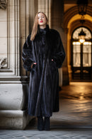 A woman wearing a long, luxurious black fur coat with a high collar and wide sleeves, standing in a grand stone corridor with warm lighting.