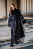 A woman wearing a long black fur coat with dark blue undertones and black boots, standing on stone steps in front of classical columns.