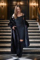 A woman stands on an ornate indoor staircase wearing a black off-the-shoulder dress, a long black faux-fur coat, and black over-the-knee boots. The setting features warm wall lighting and a polished interior.