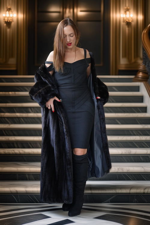 A woman stands on an ornate indoor staircase wearing a black off-the-shoulder dress, a long black faux-fur coat, and black over-the-knee boots. The setting features warm wall lighting and a polished interior.