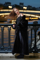 A woman posing on a bridge at night wearing a luxurious black fur coat over a black dress, with the Seine River and illuminated bridge in the background.