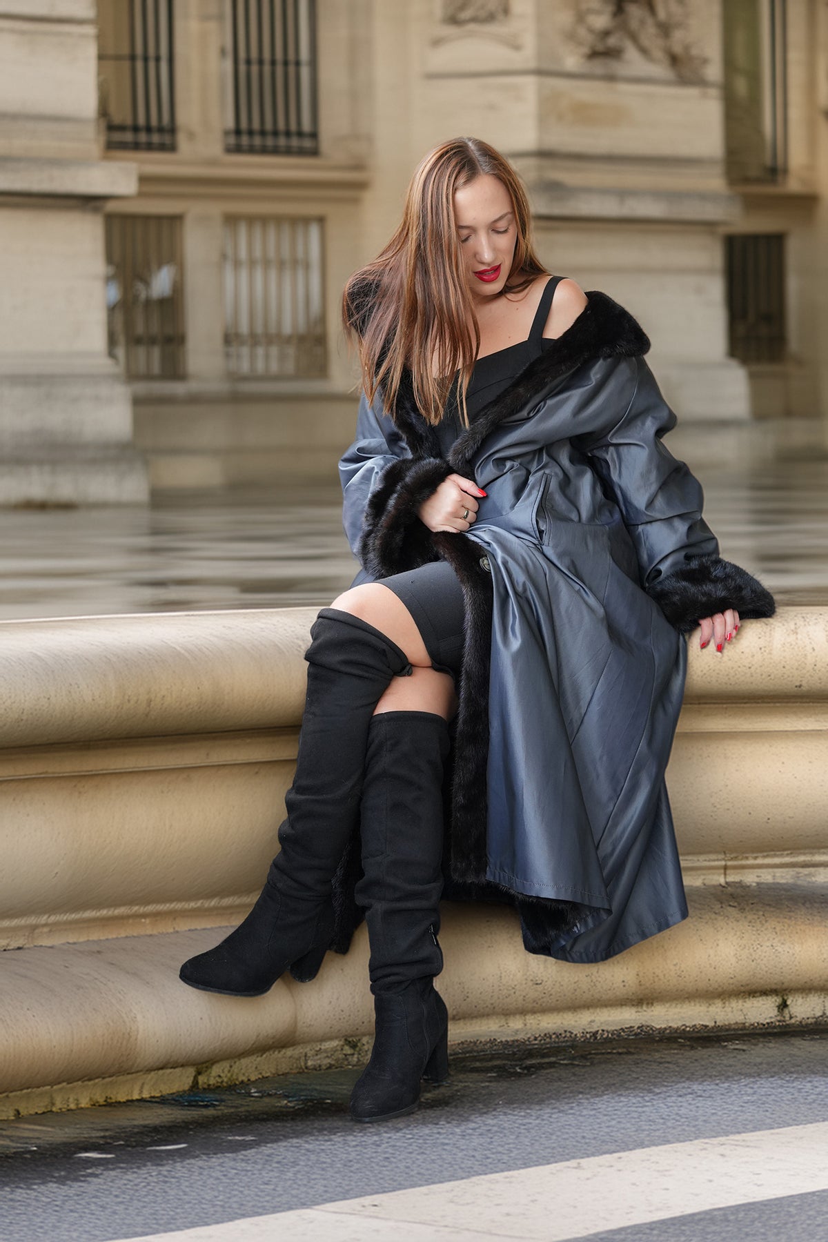 A woman sits on a stone ledge outdoors, wearing a dark metallic gray faux-leather coat with black fur trim, black thigh-high boots, and a black top.