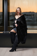 A woman sits on a windowsill wearing a luxurious black faux fur wrap, a black mini dress, and black over-the-knee boots, with a warm sunset visible through the window behind her.