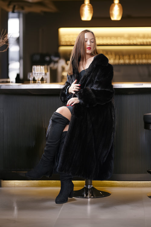 A woman sits on a bar stool in a dimly lit bar, wearing a long black fur coat over a black outfit with black over-the-knee boots.