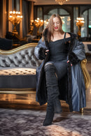 A woman models a luxurious black off-the-shoulder dress with a matching oversized satin coat featuring black fur trim, paired with black over-the-knee boots, seated on an ornate gray and gold sofa in a grand, chandelier-lit room.