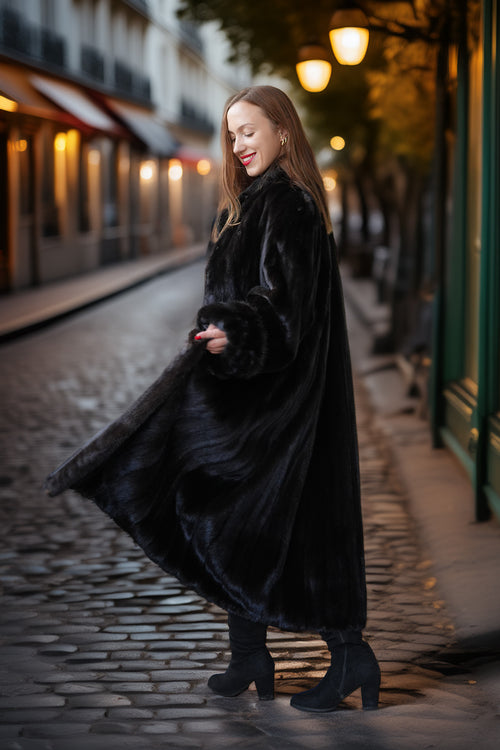 A woman wearing a long black fur coat and black ankle boots stands on a cobblestone street at night, holding out the coat to show its texture and length. The background features warm streetlights and blurred buildings.