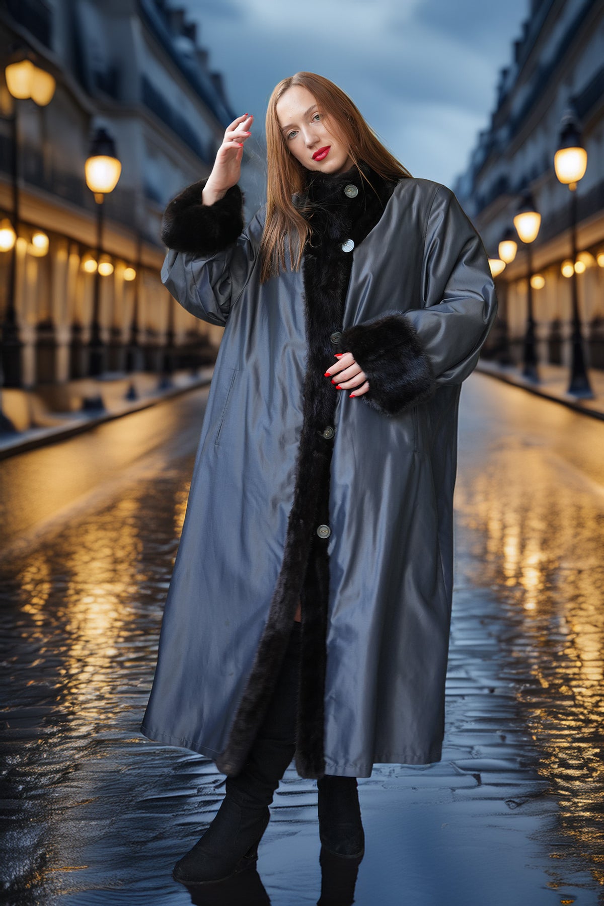A woman stands on a wet city street at dusk wearing a long dark gray coat with black fur trim over a black outfit. Street lamps glow on both sides, and her reflection is visible on the wet pavement.