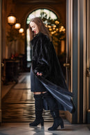 A woman wearing a long black faux fur coat over a dark dress and black over-the-knee boots, standing in a grand hallway with gold-trimmed doors and warm hanging lights.