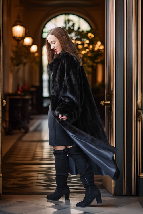 A woman wearing a long black faux fur coat over a dark dress and black over-the-knee boots, standing in a grand hallway with gold-trimmed doors and warm hanging lights.