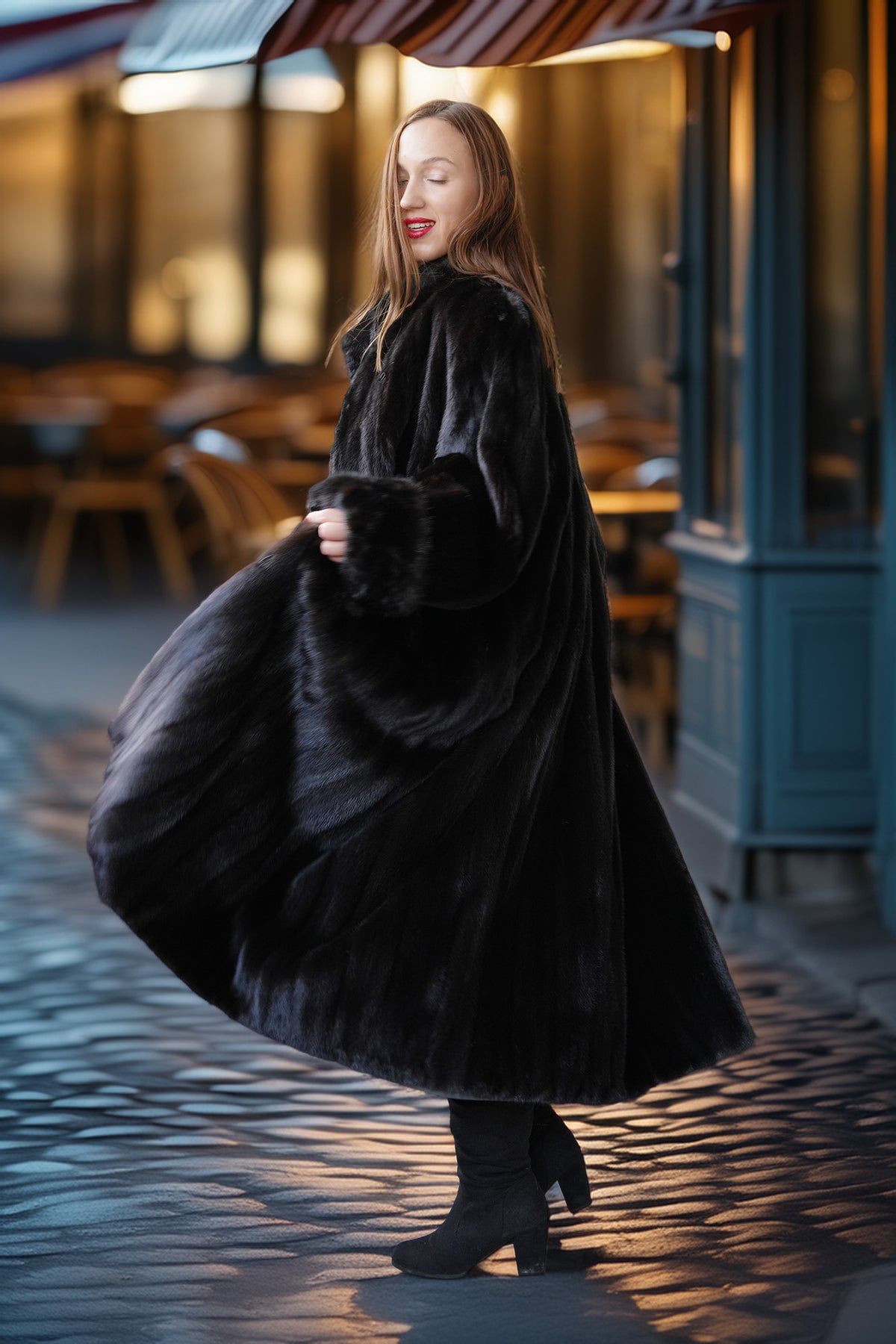 A woman wearing a long black fur coat and black boots stands on a cobblestone street in front of a cafe, holding out the side of the coat to show its texture and volume.