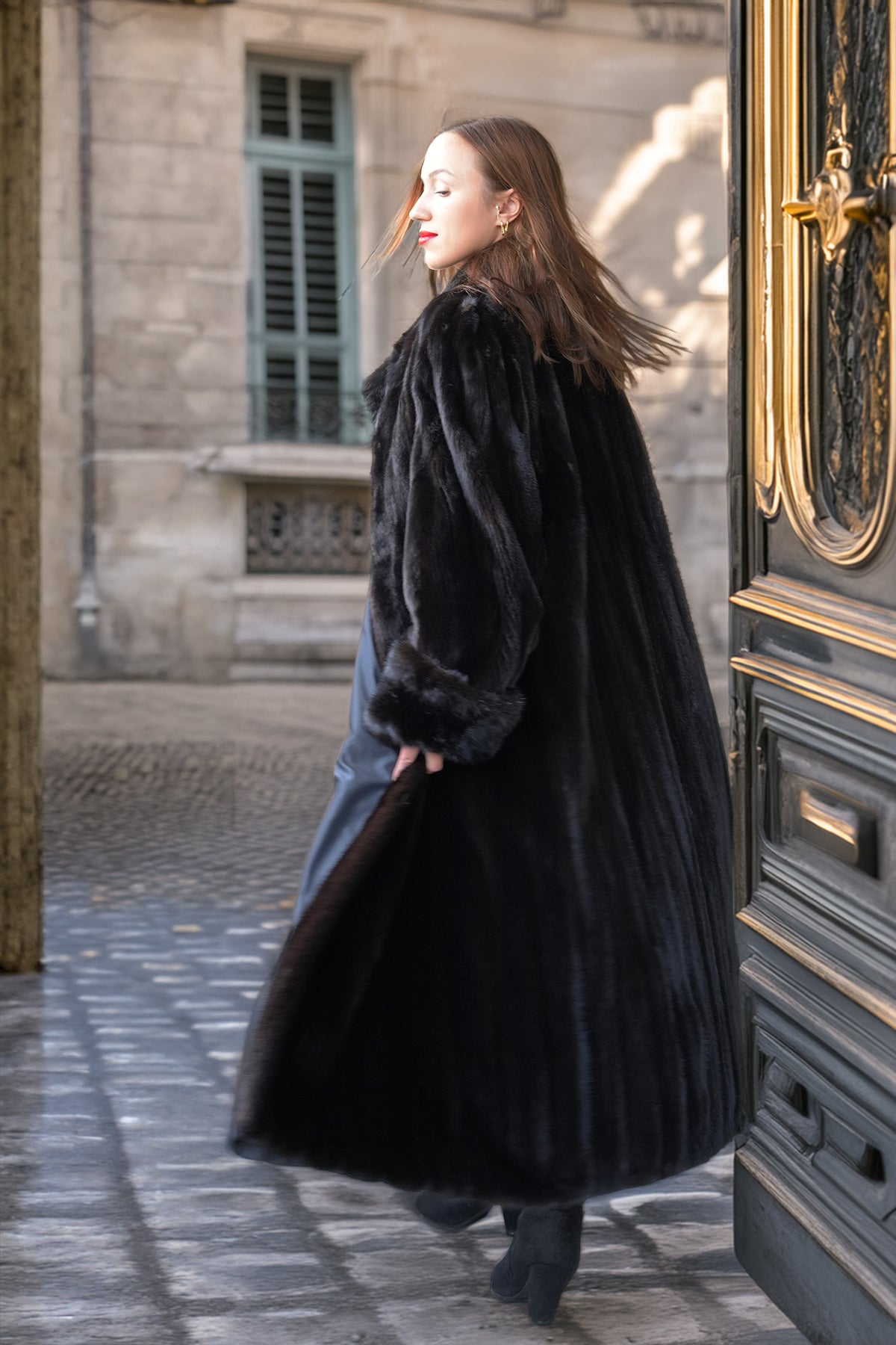 A woman wearing a long black fur coat and blue jeans walks out of a large ornate dark door onto a cobblestone street.