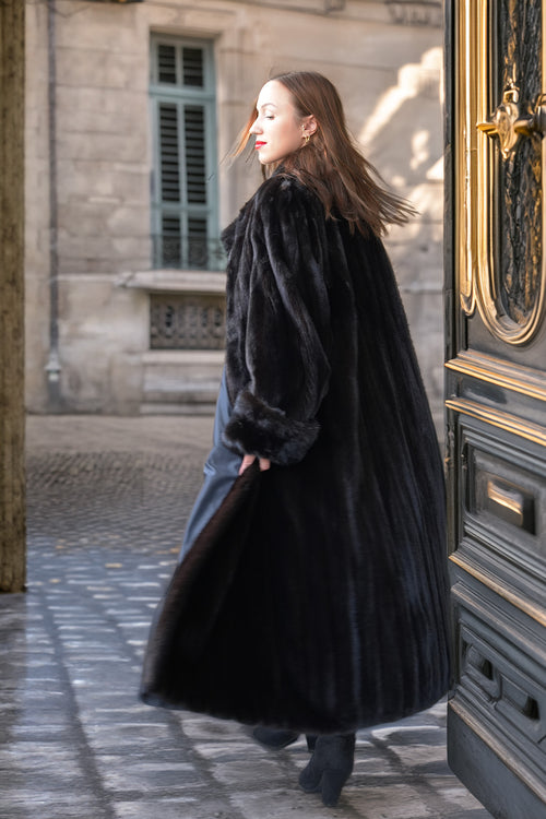 A woman wearing a long black fur coat and blue jeans walks out of a large ornate dark door onto a cobblestone street.