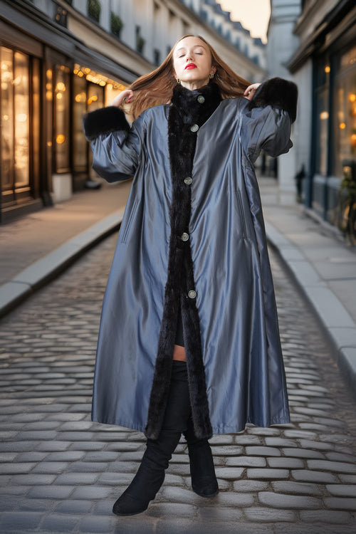 A woman stands on a cobblestone street wearing a long, shiny slate-blue coat with black faux-fur trim and a black faux-fur collar. She is wearing black knee-high boots.
