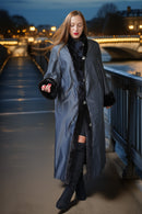 A woman wearing a long gray coat with black faux-fur trim walks on a bridge at night with city lights in the background.