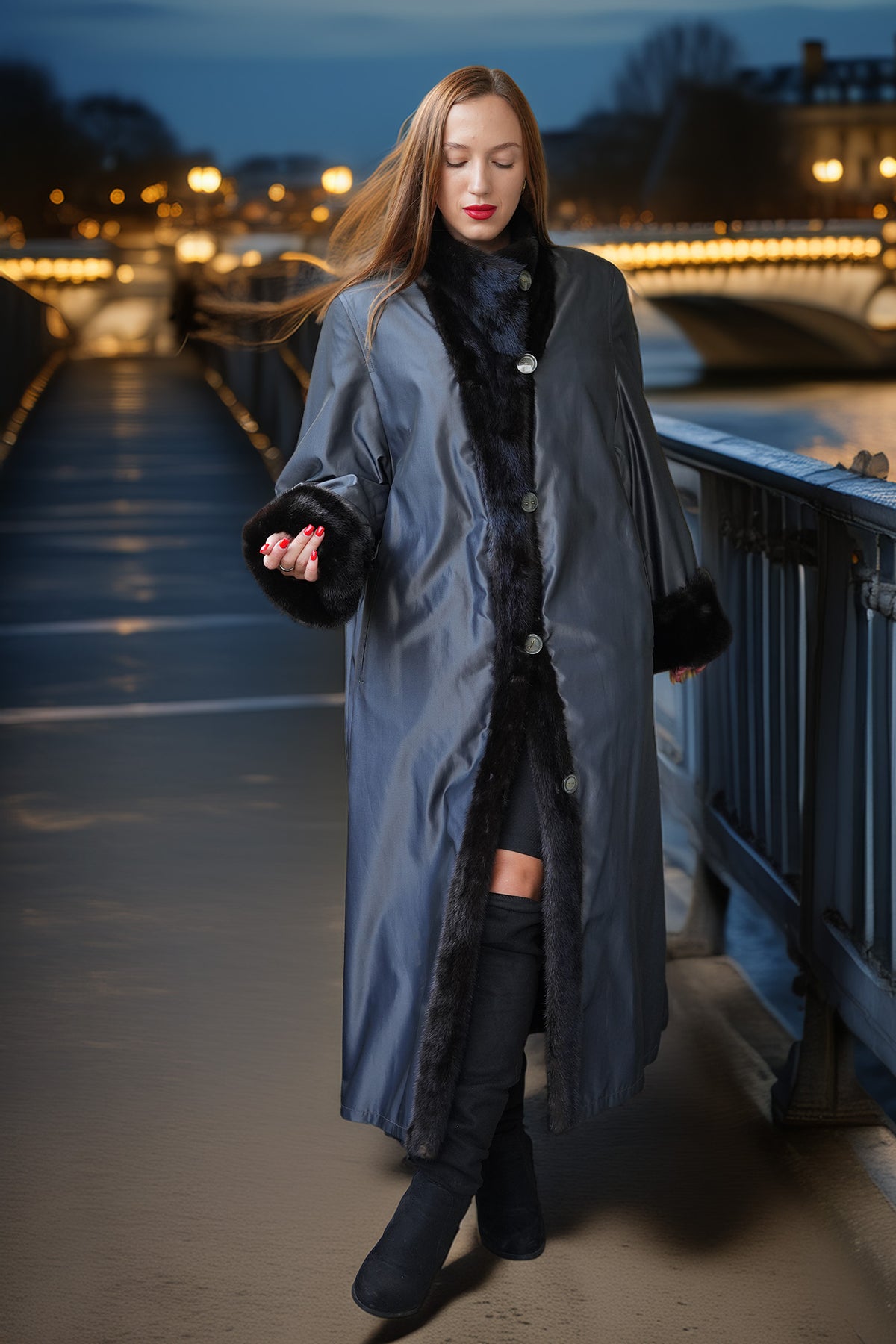 A woman wearing a long gray coat with black faux-fur trim walks on a bridge at night with city lights in the background.