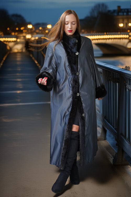A woman wearing a long gray coat with black faux-fur trim walks on a bridge at night with city lights in the background.
