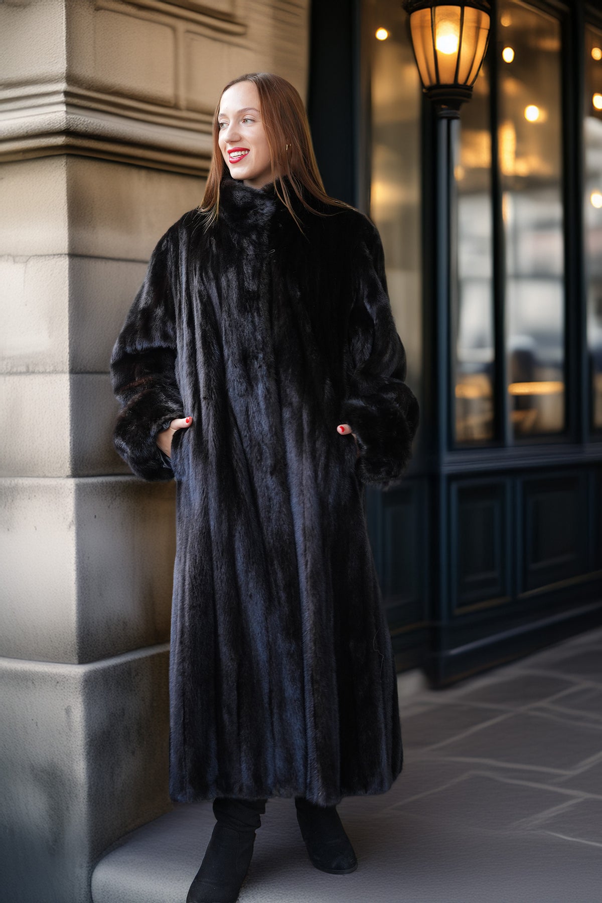 A woman wearing a long, luxurious black fur coat with a high collar and wide cuffs, standing outdoors against a stone building and dark windowed facade.