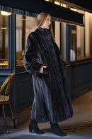 A woman wearing a long, luxurious dark charcoal gray fur coat with a high collar and wide sleeves, standing in front of a dimly lit storefront at night.