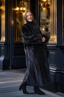 A woman wearing a luxurious black fur coat and black ankle boots stands in front of an elegant dark blue building facade with gold-trimmed windows.