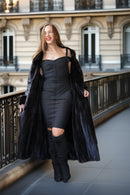 A woman stands on an outdoor balcony wearing a long black faux-fur coat over a fitted black dress and black over-the-knee boots, with a Paris-style building in the background.