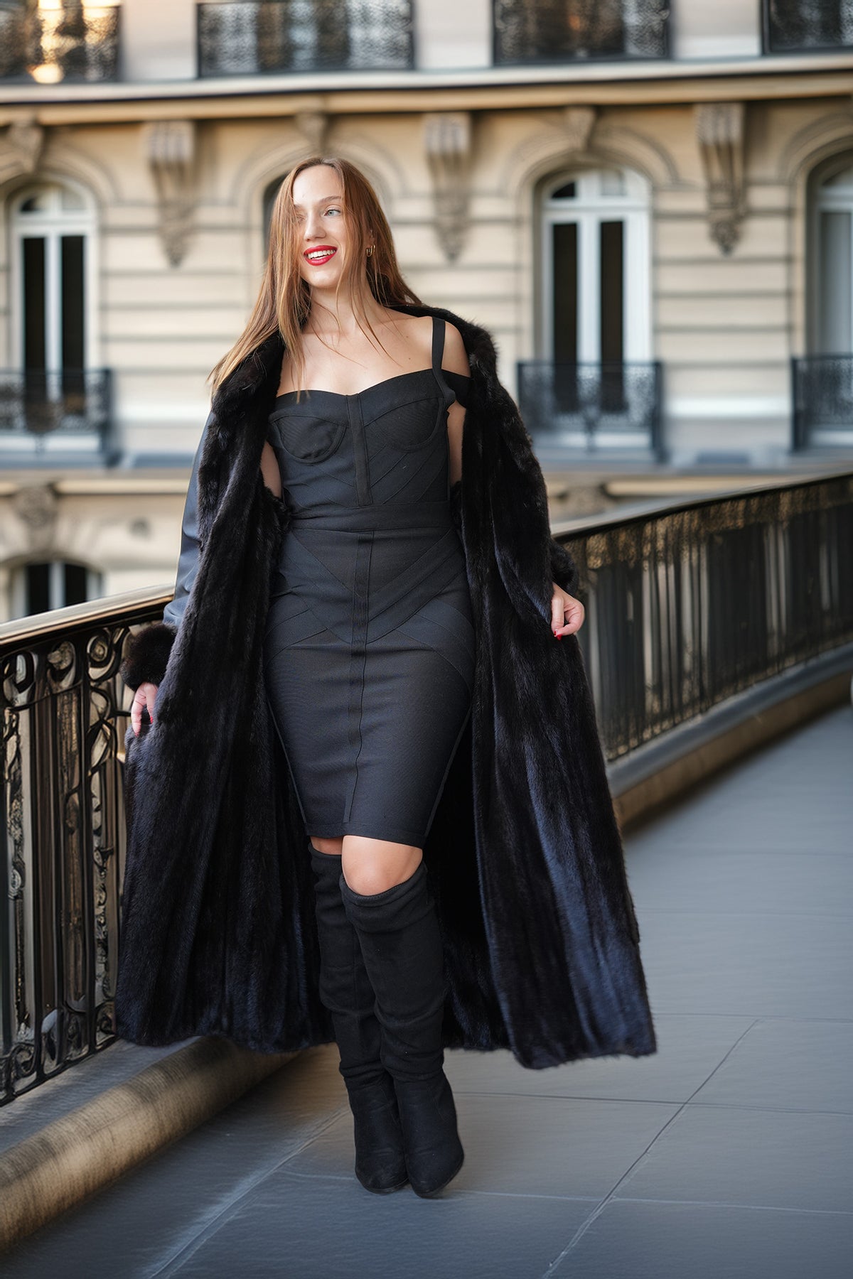 A woman stands on an outdoor balcony wearing a long black faux-fur coat over a fitted black dress and black over-the-knee boots, with a Paris-style building in the background.