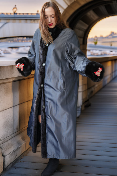 A woman wearing a long gray coat with black fur trim and black gloves, standing on a wooden walkway outdoors near a bridge at sunset.