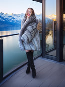 Woman in a fur coat standing on a balcony with a scenic view of mountains and water.