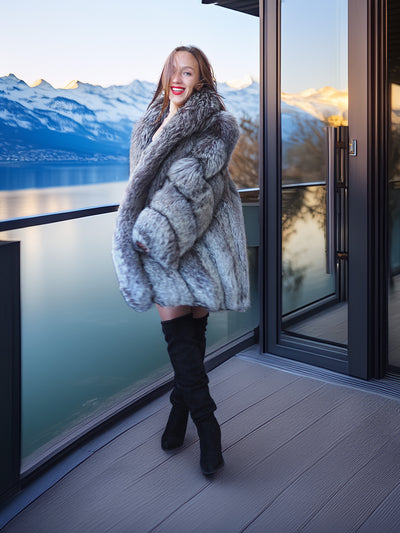 Woman in a fur coat standing on a balcony with a scenic view of mountains and water.