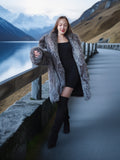 Woman in a fur coat standing by a lake with mountains in the background