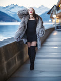 Person in a gray fur coat and black outfit standing on a wooden pier with mountains and water in the background